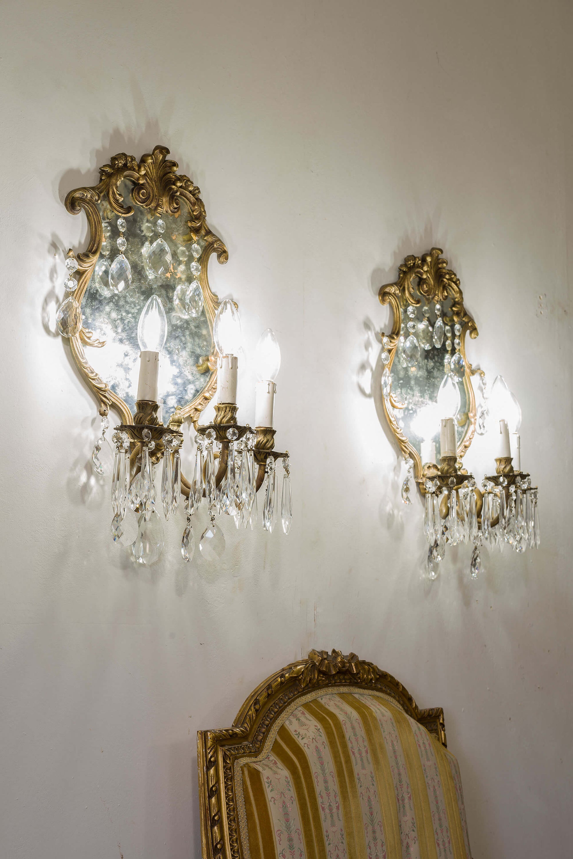 Splendida coppia di applique anni '20 con specchio centrale che amplifica la luce delle 3 lampadine E14. Realizzate in bronzo impreziosito da gocce di cristallo pendenti. Ottime condizioni generali, specchio con macchie di ossidazione argento che conferiscono un valore aggiunto del tempo trascorso. | Lo Stile Italiano