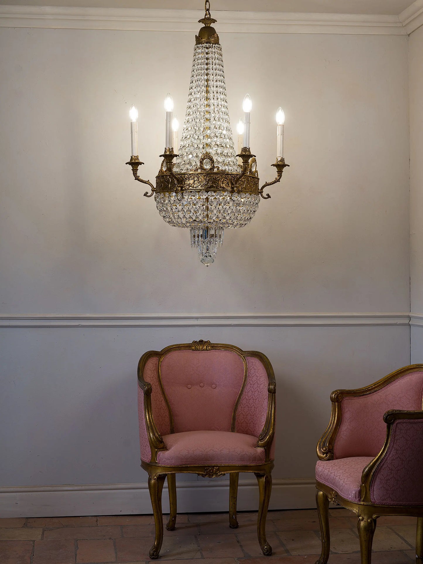Lampadario vintage stile Impero in cristallo e ottone acceso, ambientato sopra poltrone classiche in un interno elegante | Lo Stile Italiano